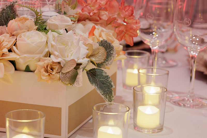 A floral centerpiece with white and peach flowers next to votive candles and empty wine glasses on a table.