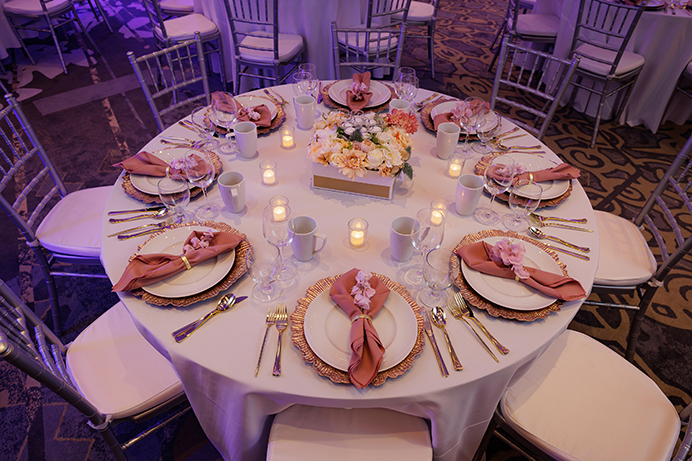 Round table set for eight with pink napkins, floral centerpiece, candles, and elegant tableware at a formal event.