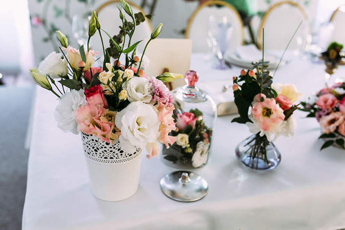 A white table with floral arrangements in vases and a glass cloche covering small flowers, set for an event.