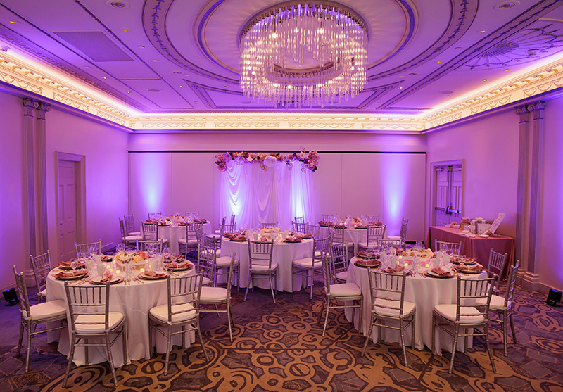 A banquet hall with round tables set for an event, purple uplighting, a chandelier, and a floral arch at the front.